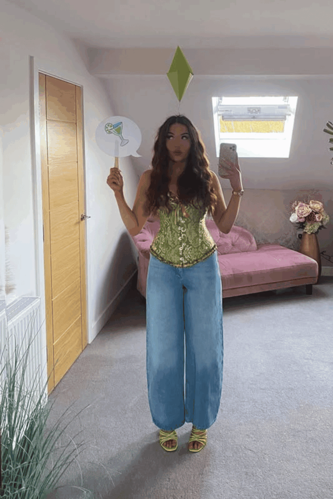Woman dressed as a Sims character with a green plumbob headpiece and a thought bubble prop, taking a mirror selfie.