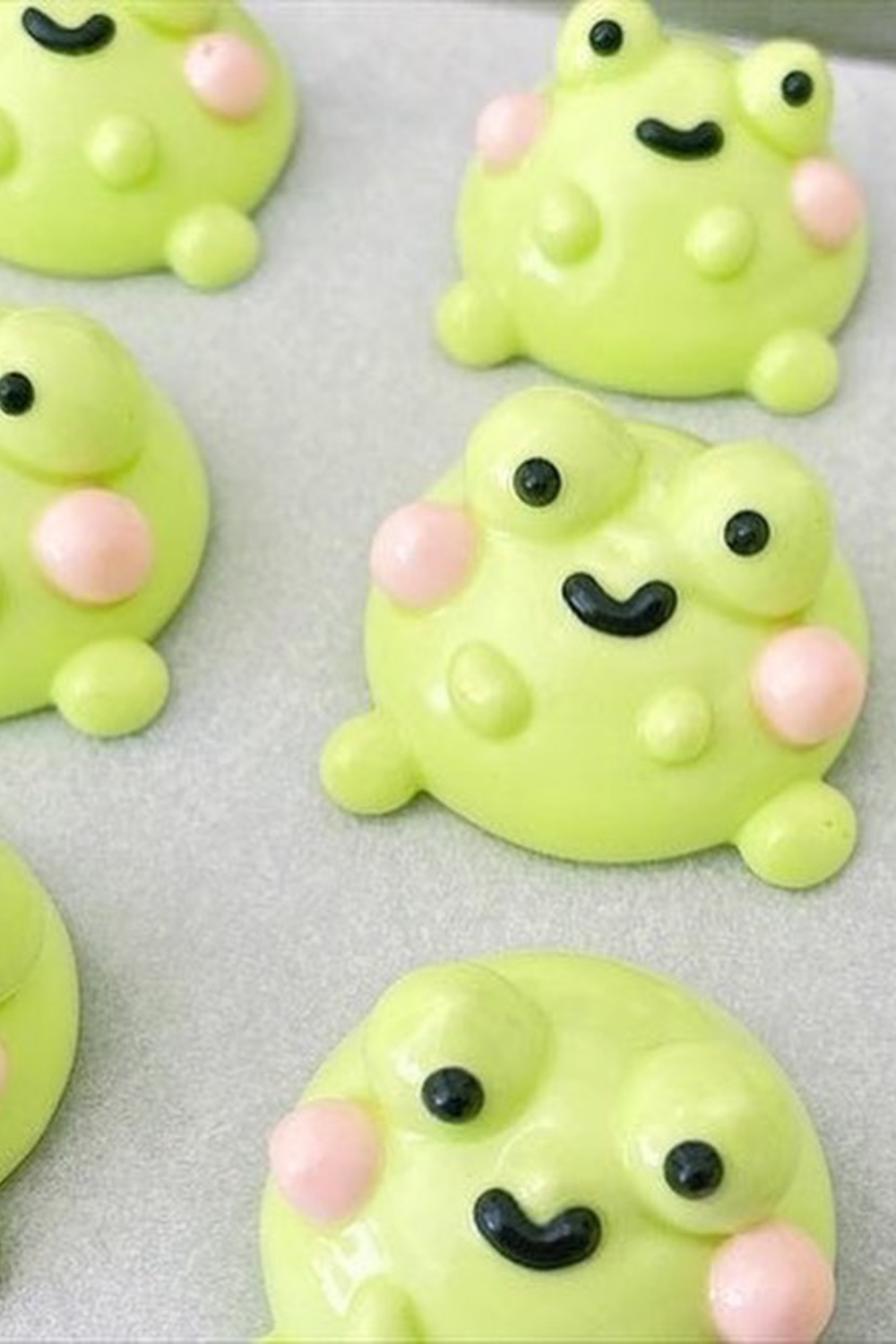 Bright green meringue cookies shaped like frogs, featuring blushing cheeks and happy expressions, arranged on a baking sheet.