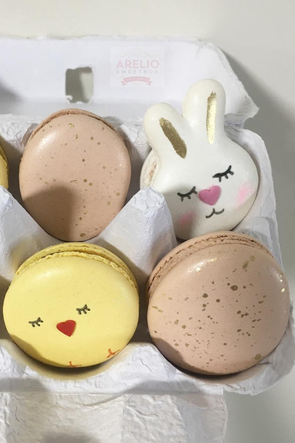 A carton of pastel-colored macarons designed as bunnies and chicks with delicate golden accents.