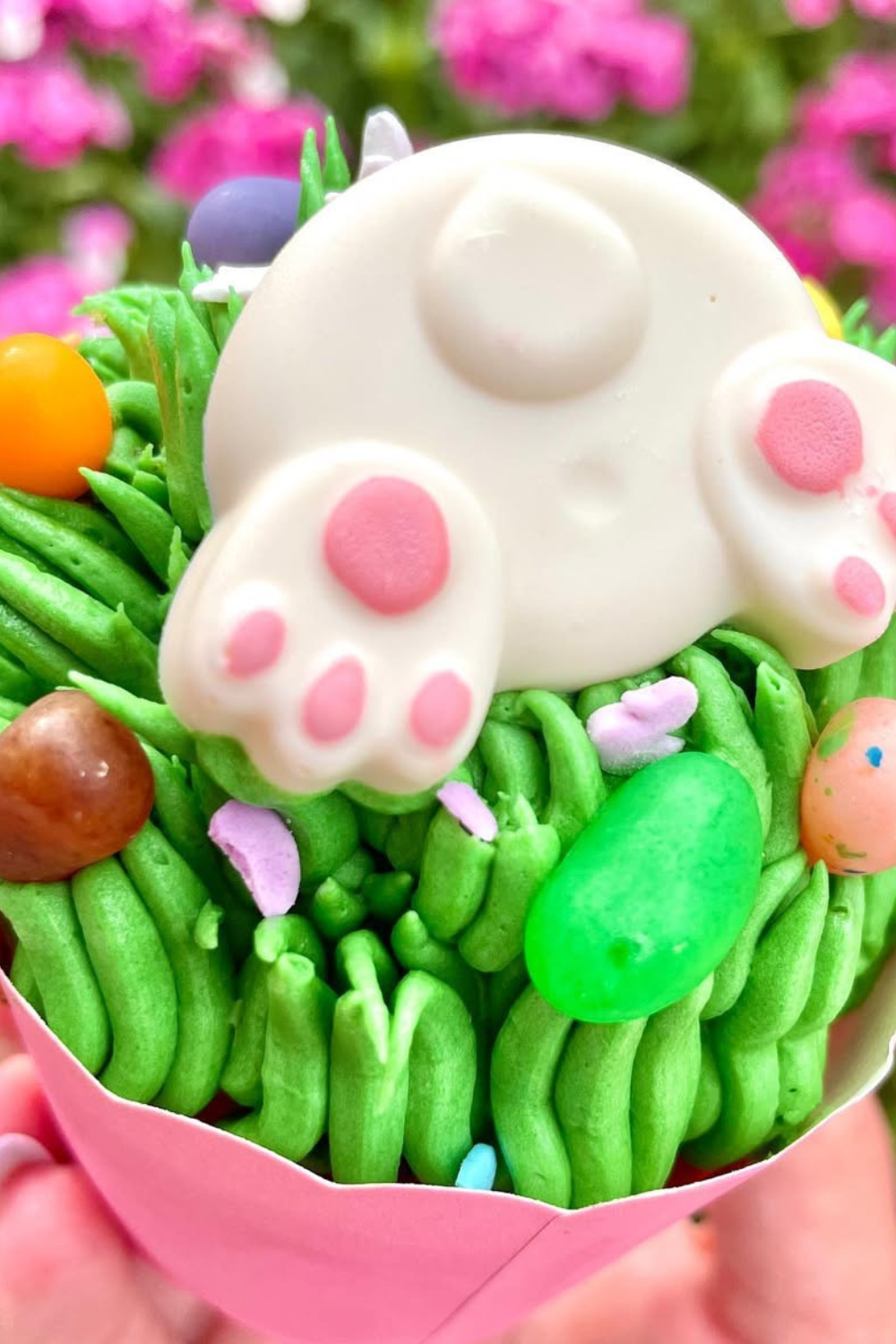 A cupcake topped with green frosting "grass," colorful candies, and a bunny bottom decoration with pink paws.