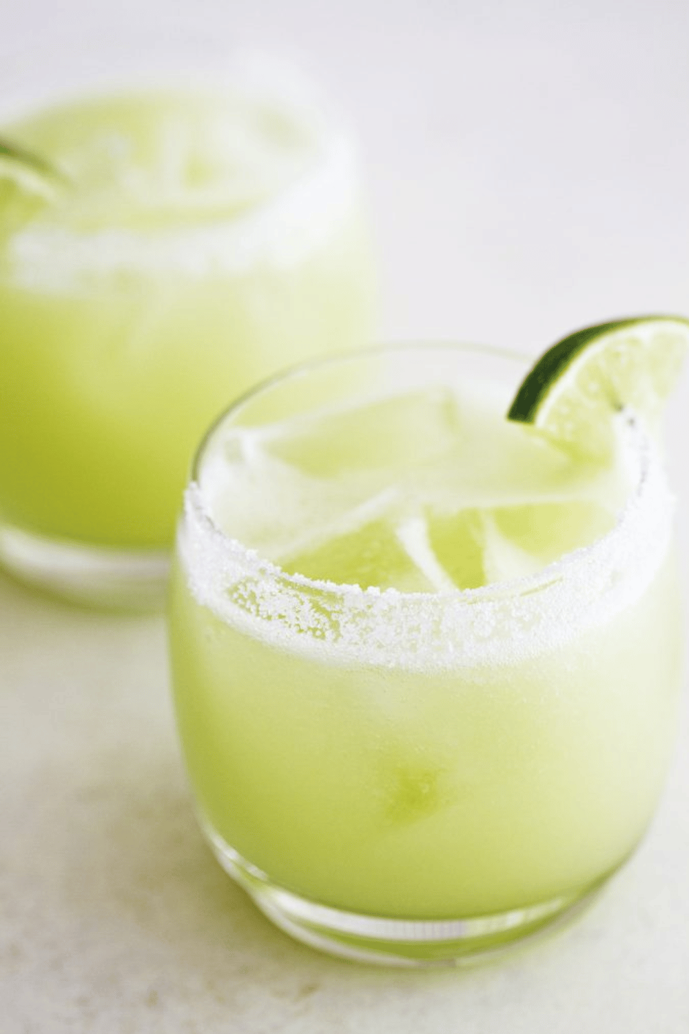 Two honeydew lime margaritas in salt-rimmed glasses with lime wedges.