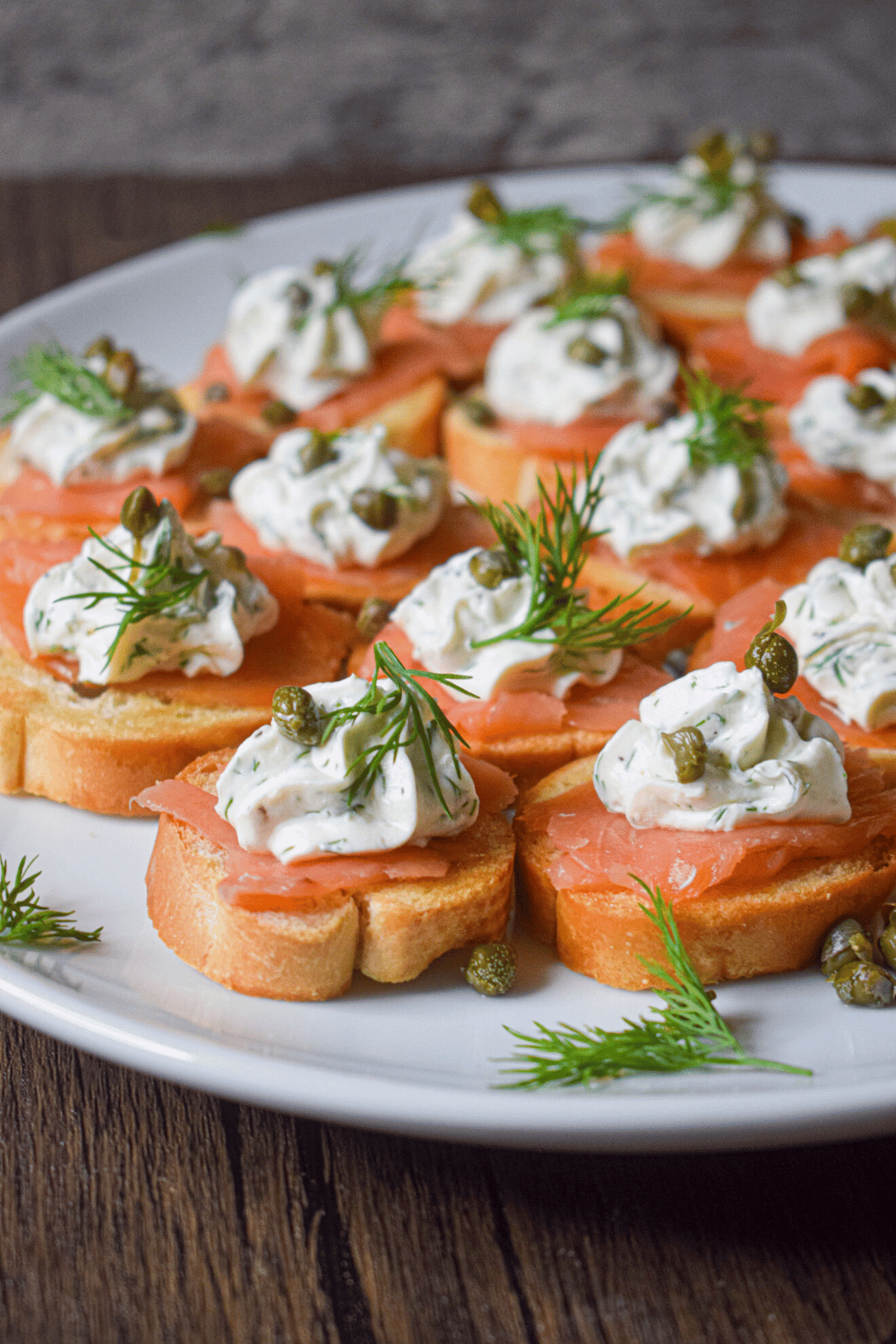Smoked salmon crostini with herbed cream, fresh dill, and capers on a white serving platter.