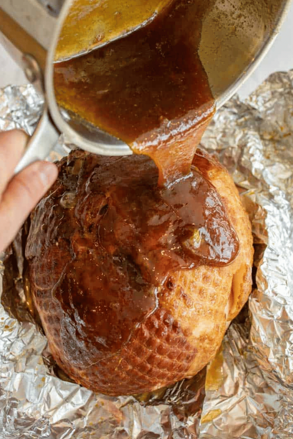 A whole ham being glazed with brown sugar sauce on foil.