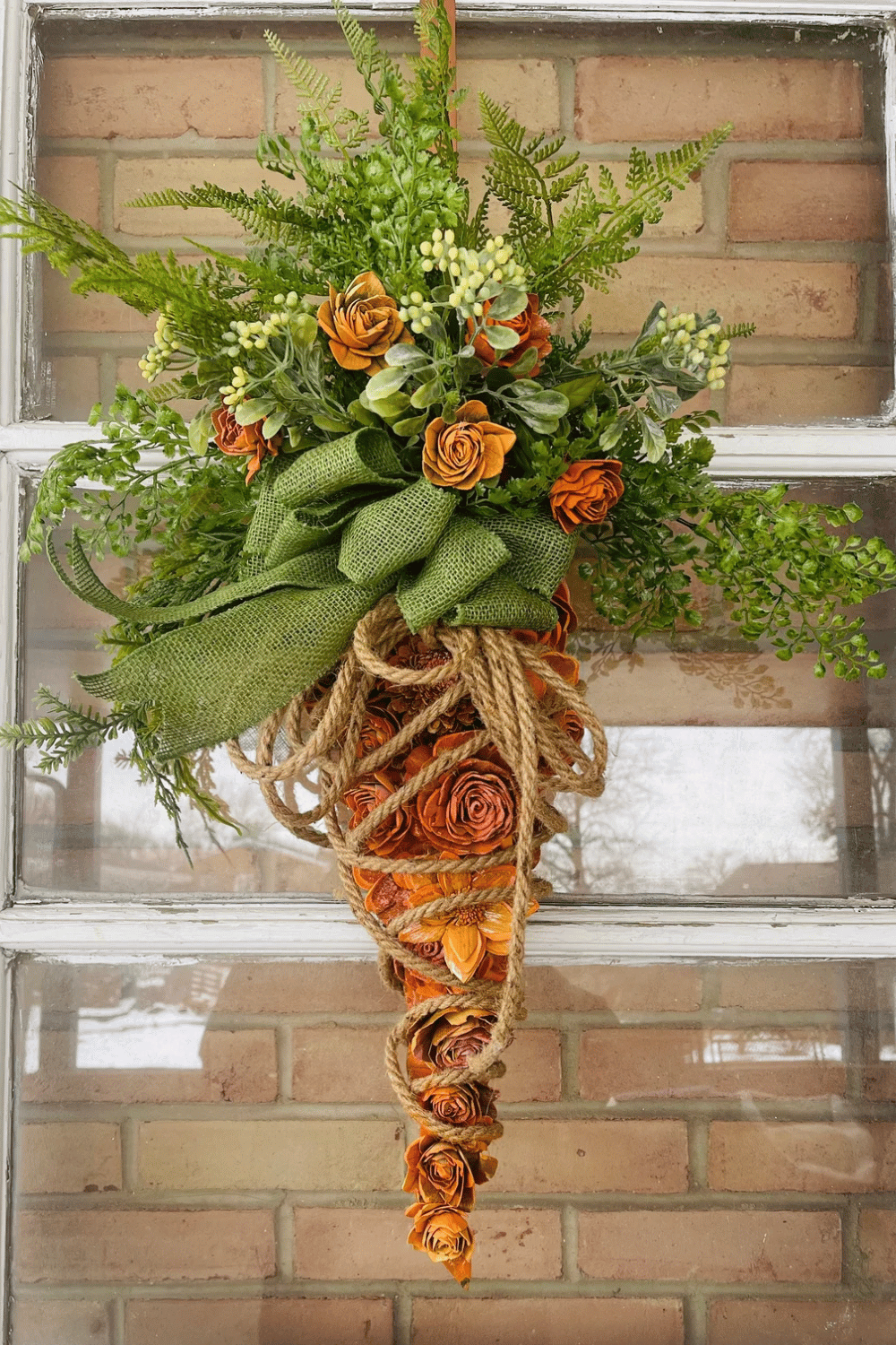 A homey swag wreath takes the shape of a carrot and features greenery and orange flowers.