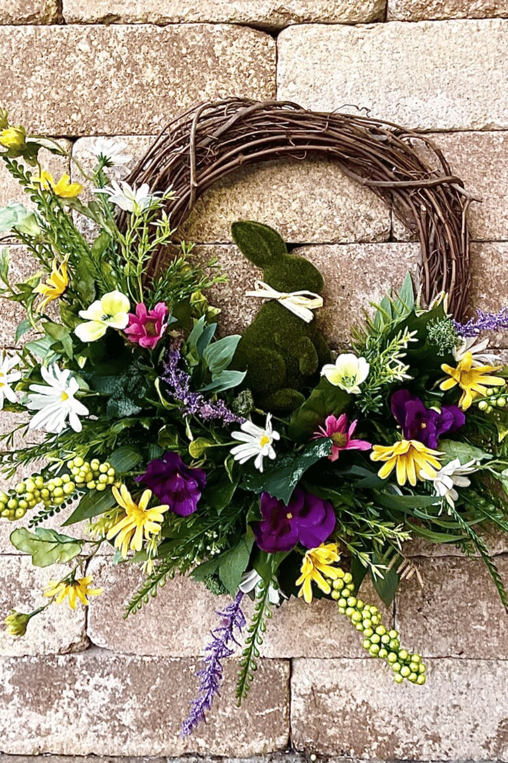 A grapevine base supports an Easter wreath which features a moss bunny among wildflowers.
