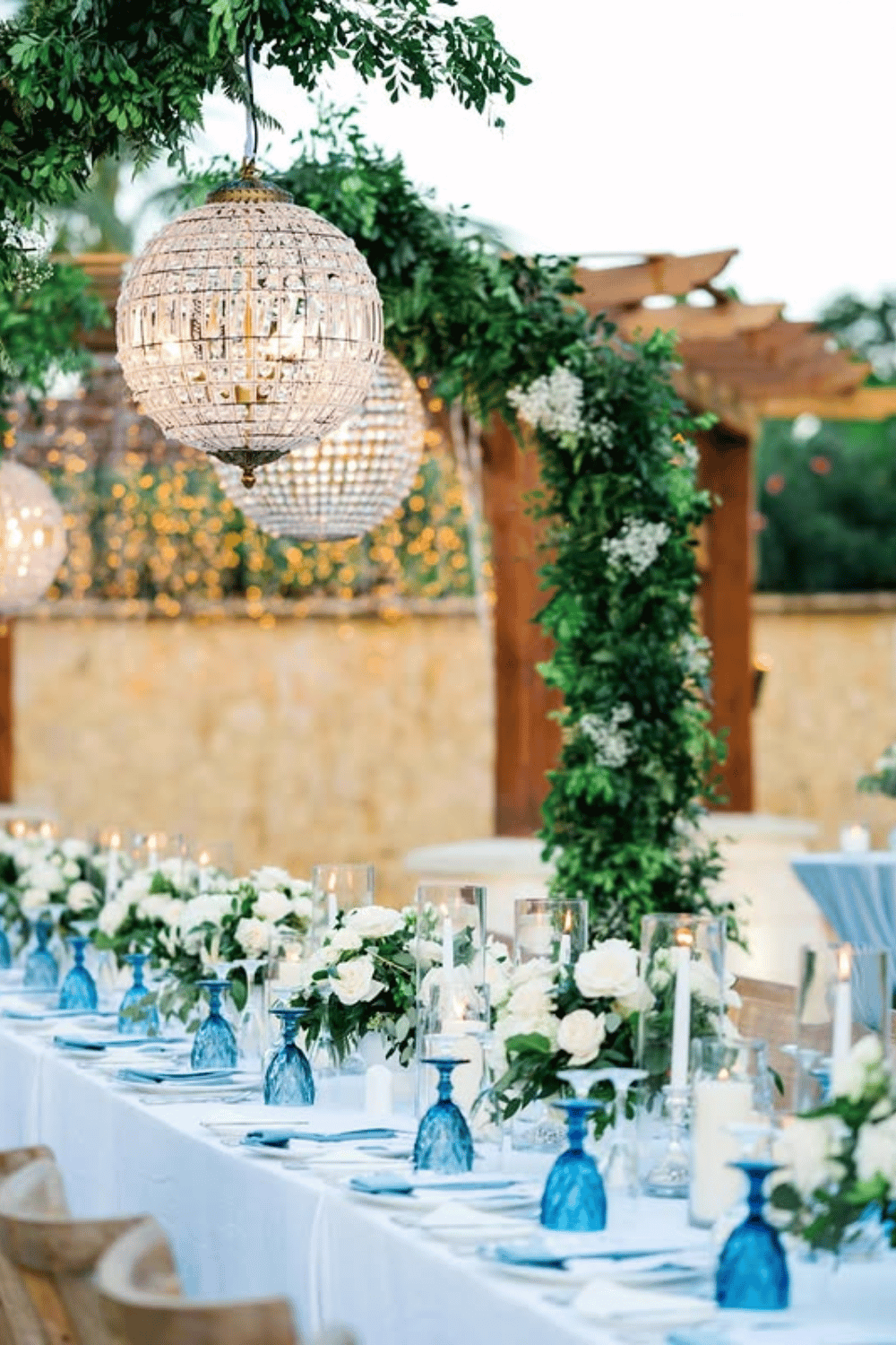 Long coastal wedding table with blue glassware, greenery, and crystal chandeliers. Wedding Table Styling
