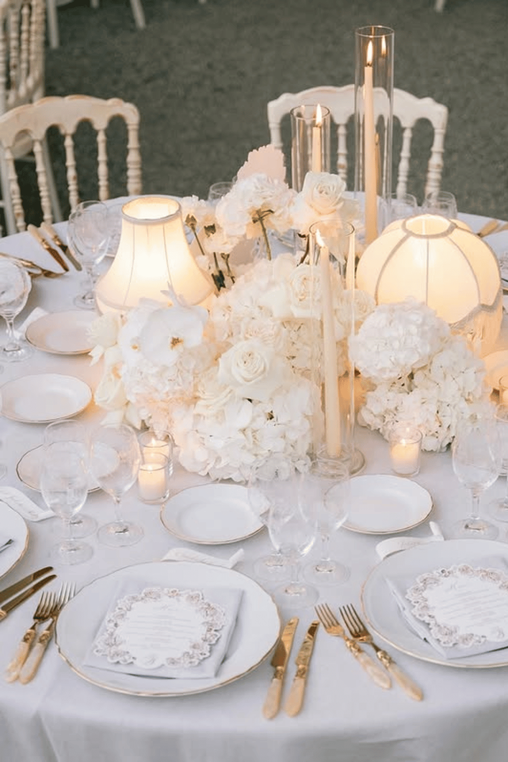 Elegant all-white table setting with gold flatware and glowing lamps for wedding reception