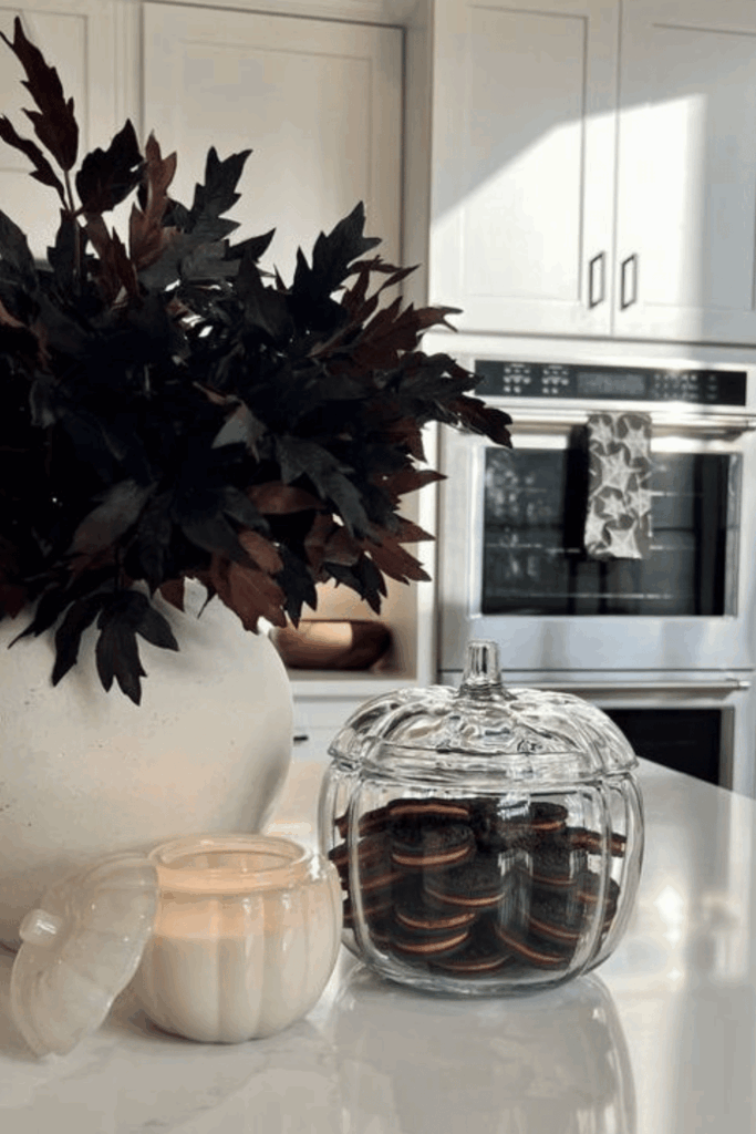 Modern white kitchen with glass pumpkin jar filled with cookies, candle, and dark fall leaves